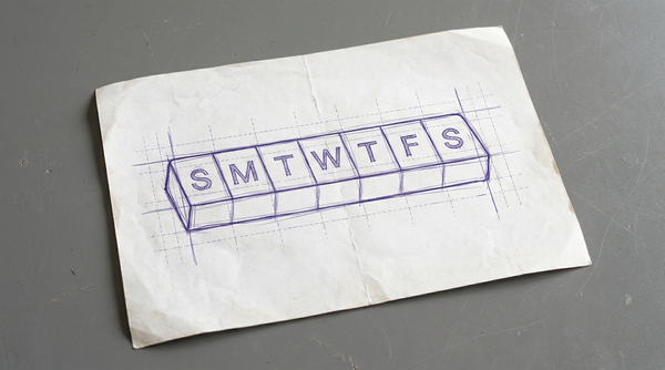 a rectangular pill organizer box with its lid closed, the top surface divided into seven distinct, equal sections, each marked with the first letter of a day of the week (S, M, T, W, T, F, S)